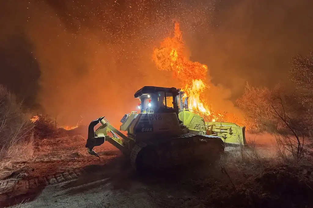 Maquinaria pesada en combate contra incendio forestal con una bulldozer en acción en un escenario nocturno, con llamas y humo intenso.